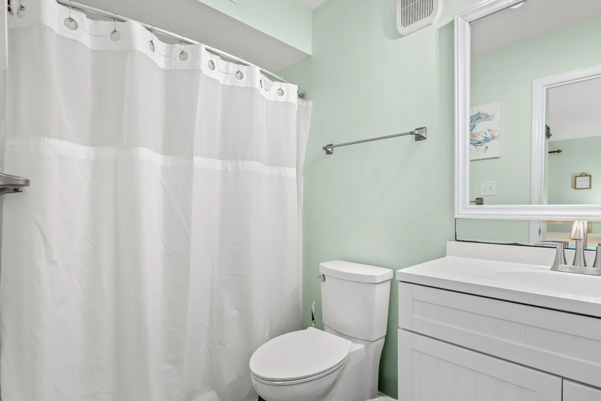 A modern bathroom with light green walls, a white shower curtain, a sleek vanity and sink, and a toilet. The space feels clean, fresh, and well-appointed, ideal for morning or evening routines.