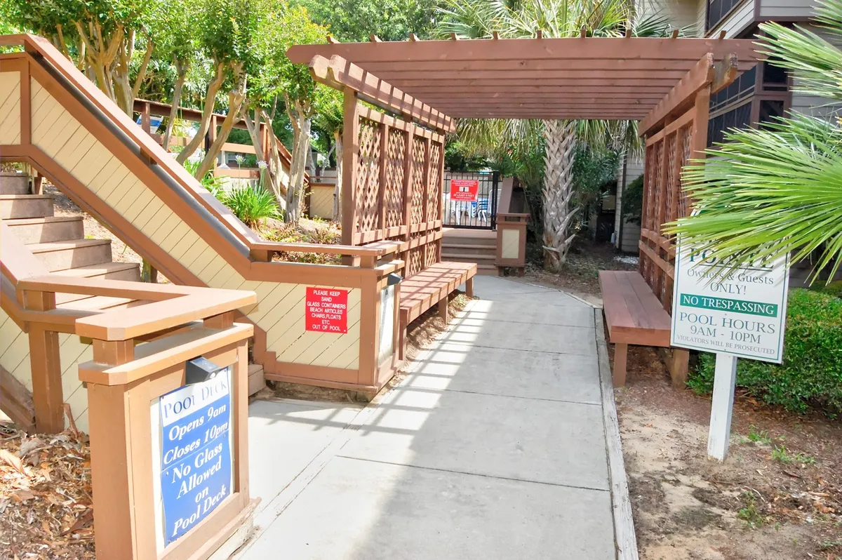 This image features a welcoming outdoor pool area framed by a wooden pergola, visible signage, and ample spots for lounging, socializing, and soaking up the sun. The pool setting exudes a relaxing, resort-inspired atmosphere ideal for both recreation and downtime with friends or family.