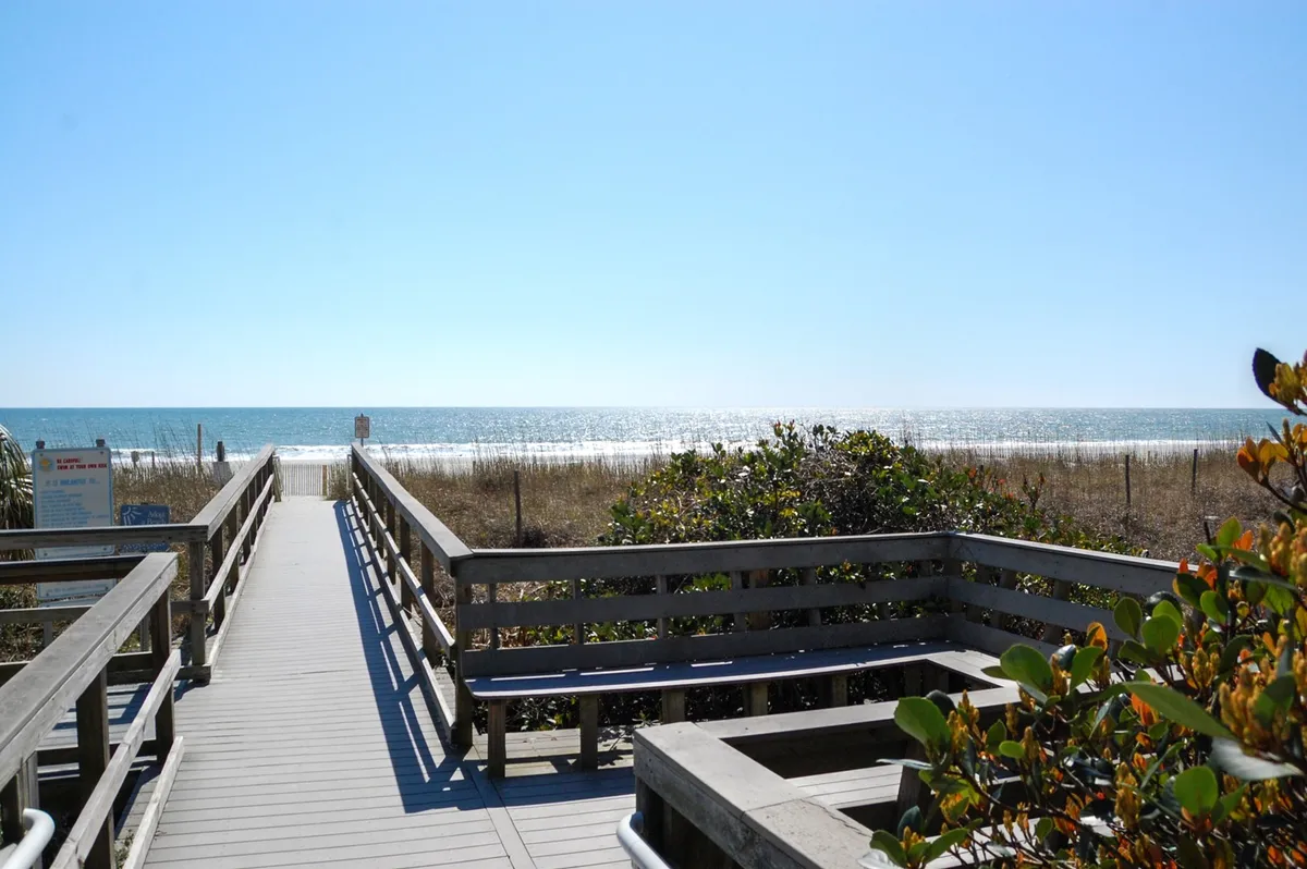 A scenic boardwalk winds through lush dunes and coastal vegetation, leading directly to the sandy beach. The ocean glimmers in the distance, inviting visitors to stroll or relax along the shoreline. The soft light of sunset adds a peaceful, golden glow.