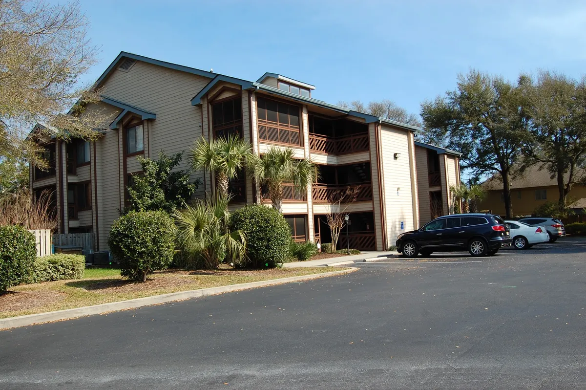 This exterior shot captures the facade of the Heron Pointe building with inviting balconies, palm trees, and a relaxed parking area. The view emphasizes the sense of community and outdoor space ideal for long-term guests.