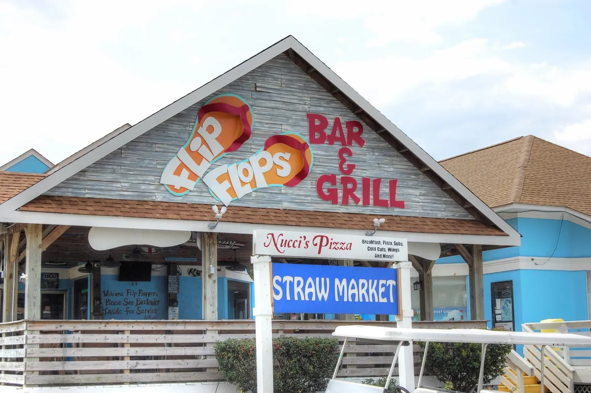 This image shows the exterior of a local dining and shopping area, featuring prominent signage for Flip Flops Bar & Grill, the Straw Market, and Nucci's Pizza. The cheerful outdoor setting suggests a lively atmosphere perfect for exploring new flavors, relaxing with a drink, or browsing through shops together.