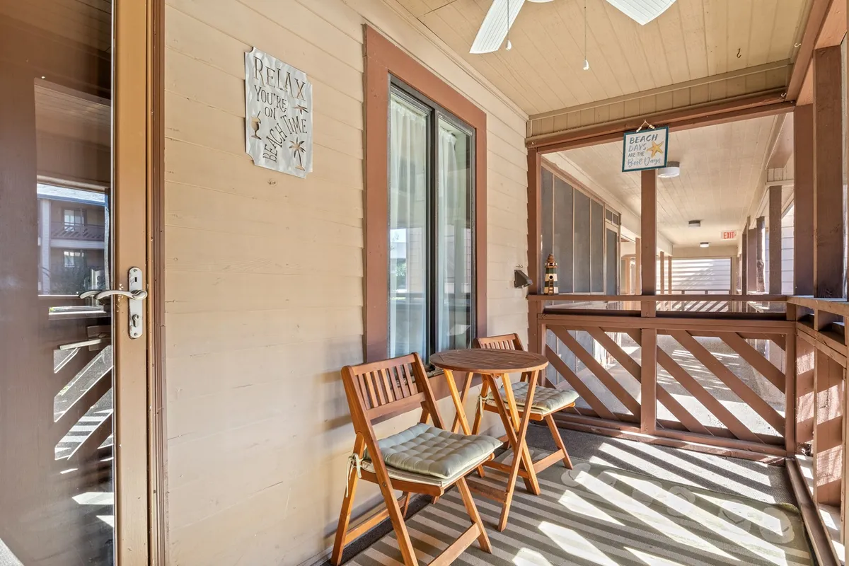 A first-floor screened porch with two wooden chairs, a small round table, ceiling fan, and a decorative sign. The space is perfectly suited for enjoying morning coffee, relaxing in the breeze, or starting the day with casual conversations among friends or family.