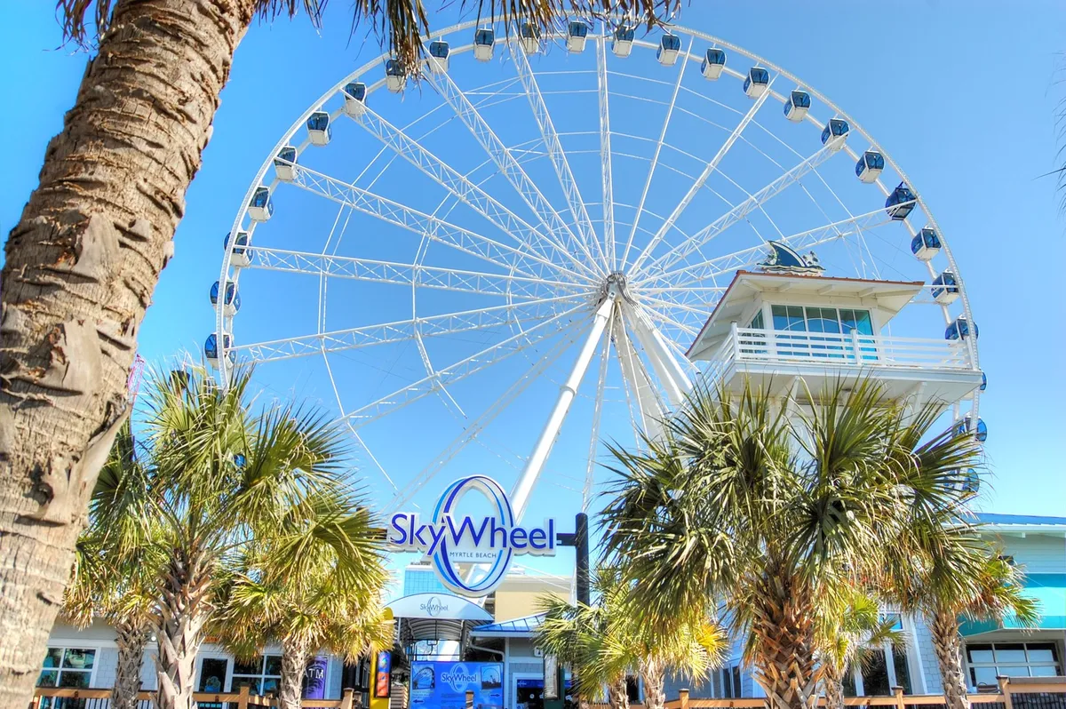 The towering SkyWheel stands against blue skies, flanked by swaying palm trees and welcoming entrance signage. Families and friends gather to enjoy panoramic views and take photos as part of their Myrtle Beach adventure.