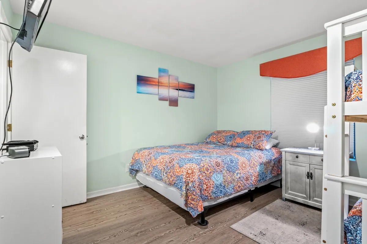 A bright, inviting bedroom with a cheerful blend of colorful bedding on a main bed and a bunk bed set against the wall. Artwork adds a playful touch, and sunlight filters in through curtains, creating a welcoming space that appeals to both families and groups of friends.