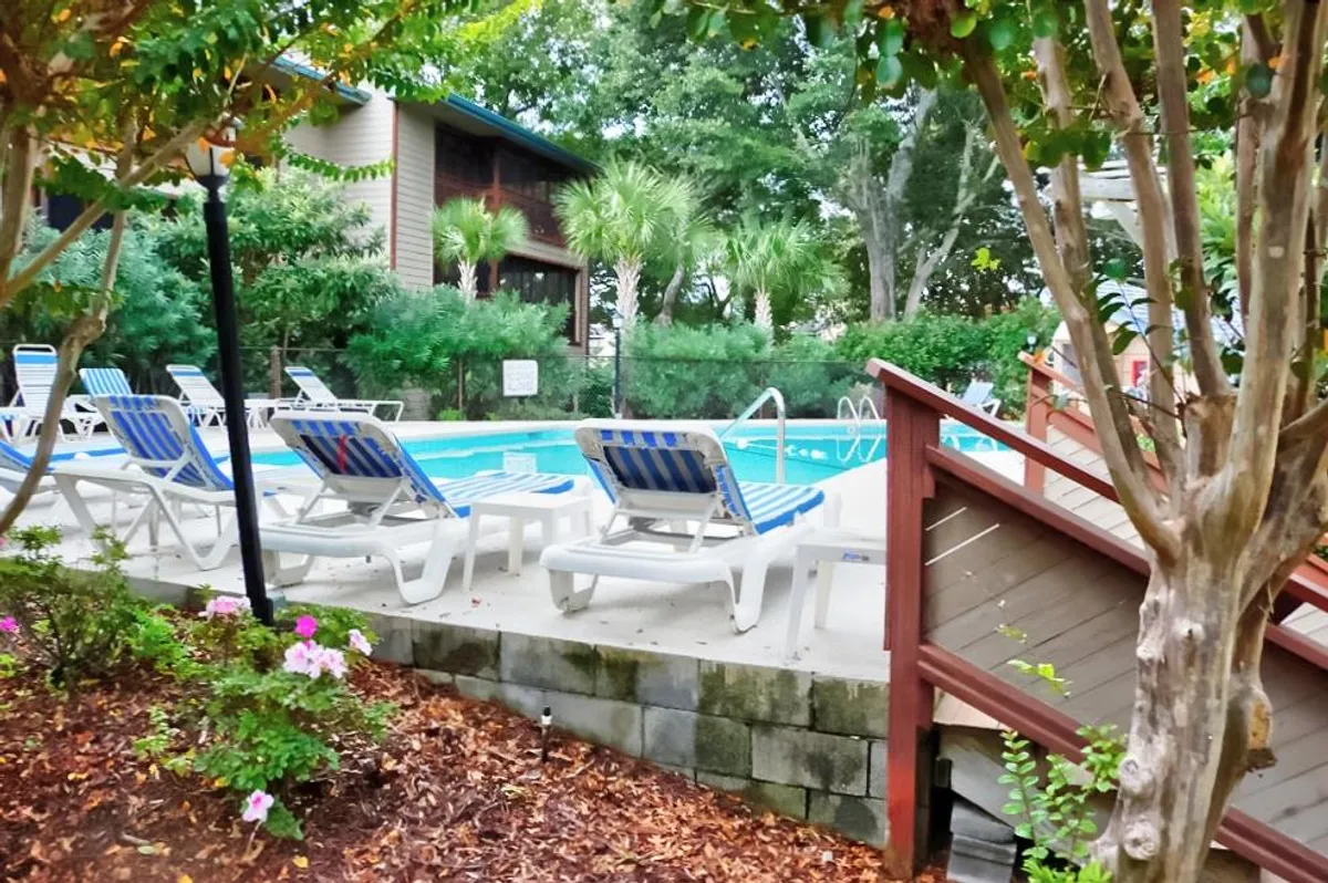 The image showcases the inviting outdoor pool area of the Heron Pointe condo complex, surrounded by lush greenery and dotted with comfortable lounge chairs. The scene exudes relaxation and leisure, with guests enjoying the pool, sunbathing, and socializing under the warm Myrtle Beach sun.