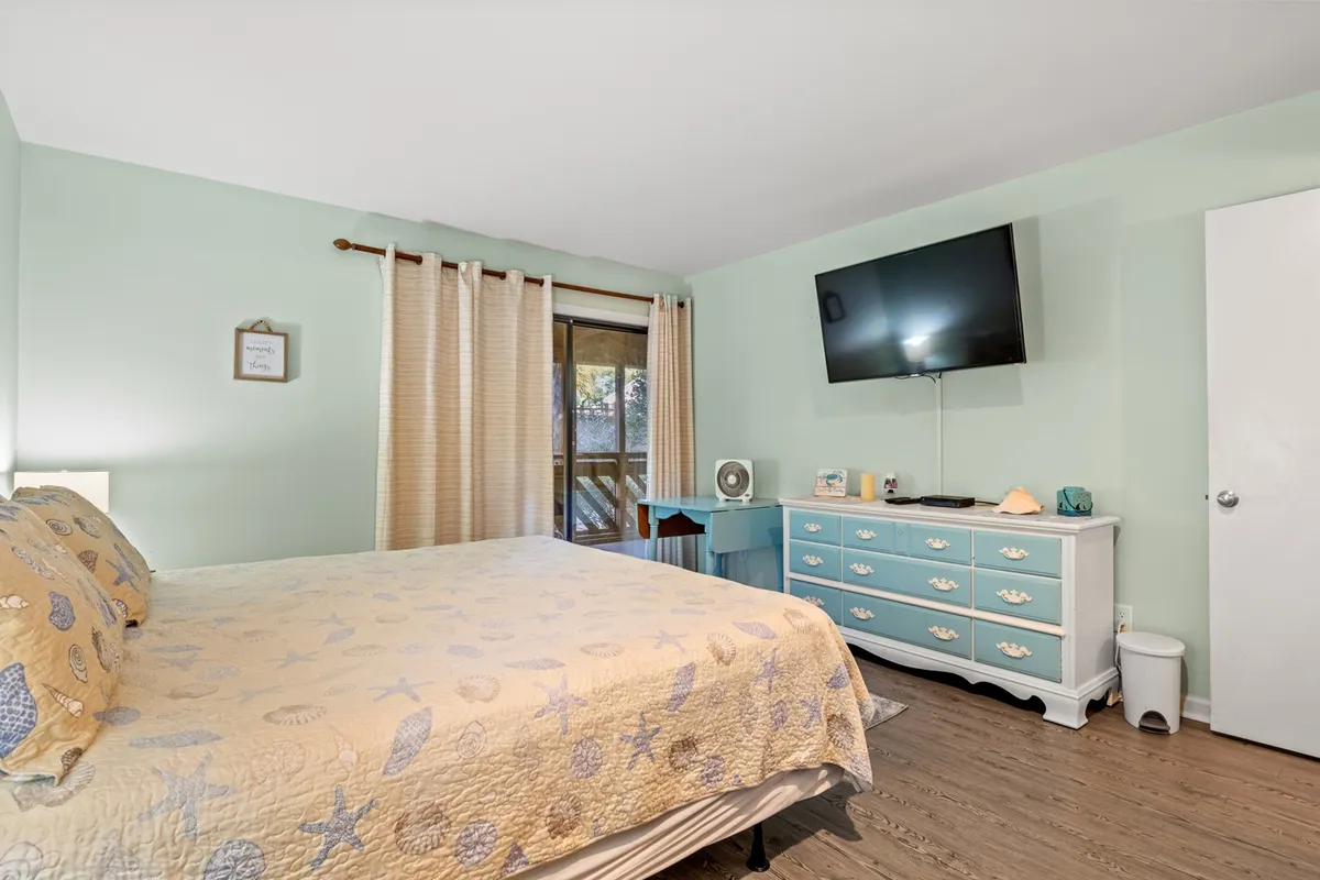 A serene bedroom featuring a bed with a soft seashell-patterned quilt, a light blue dresser, large window with drapes letting in natural light, and a wall-mounted TV. The space feels calm and restful, ideal for unwinding after busy days.