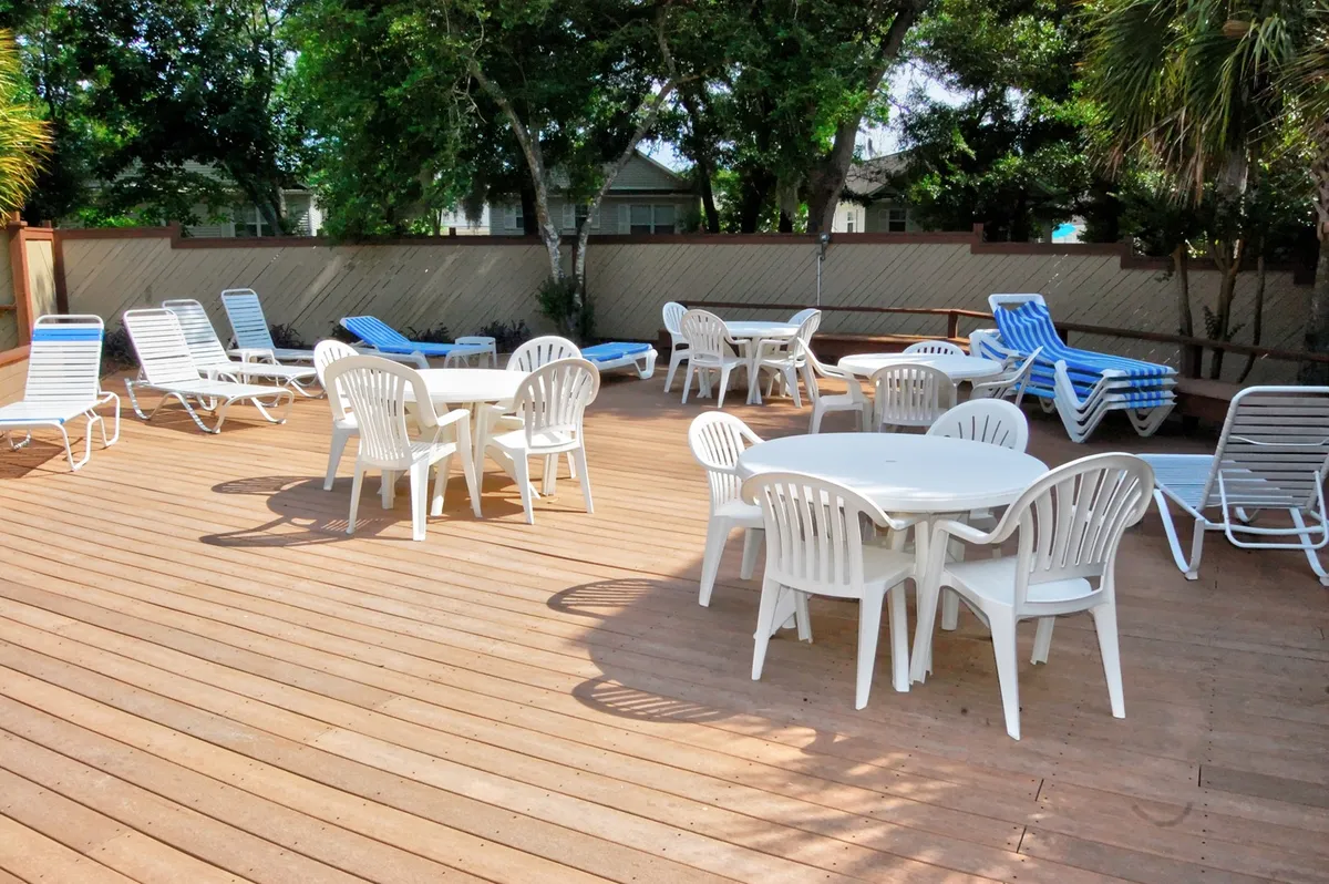 Here, white plastic tables and lounge chairs are scattered beneath the shade of mature trees, creating an inviting sun deck or gathering spot. The scene suggests moments of sunbathing, casual meals, and leisure time with others in a tranquil, resort-like setting.