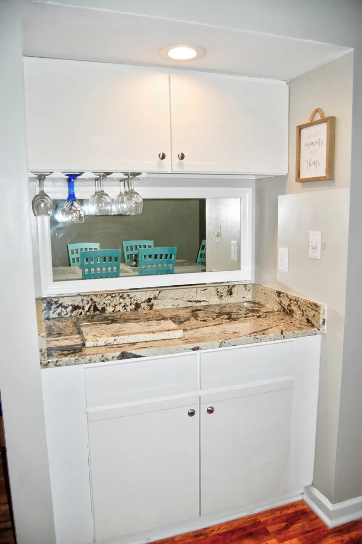 A granite countertop bar with hanging wine glasses overhead, stylish wall decor, and teal dining chairs reflected in a mirror. The setup suggests a space ideal for entertaining, preparing drinks, and hosting memorable meals.